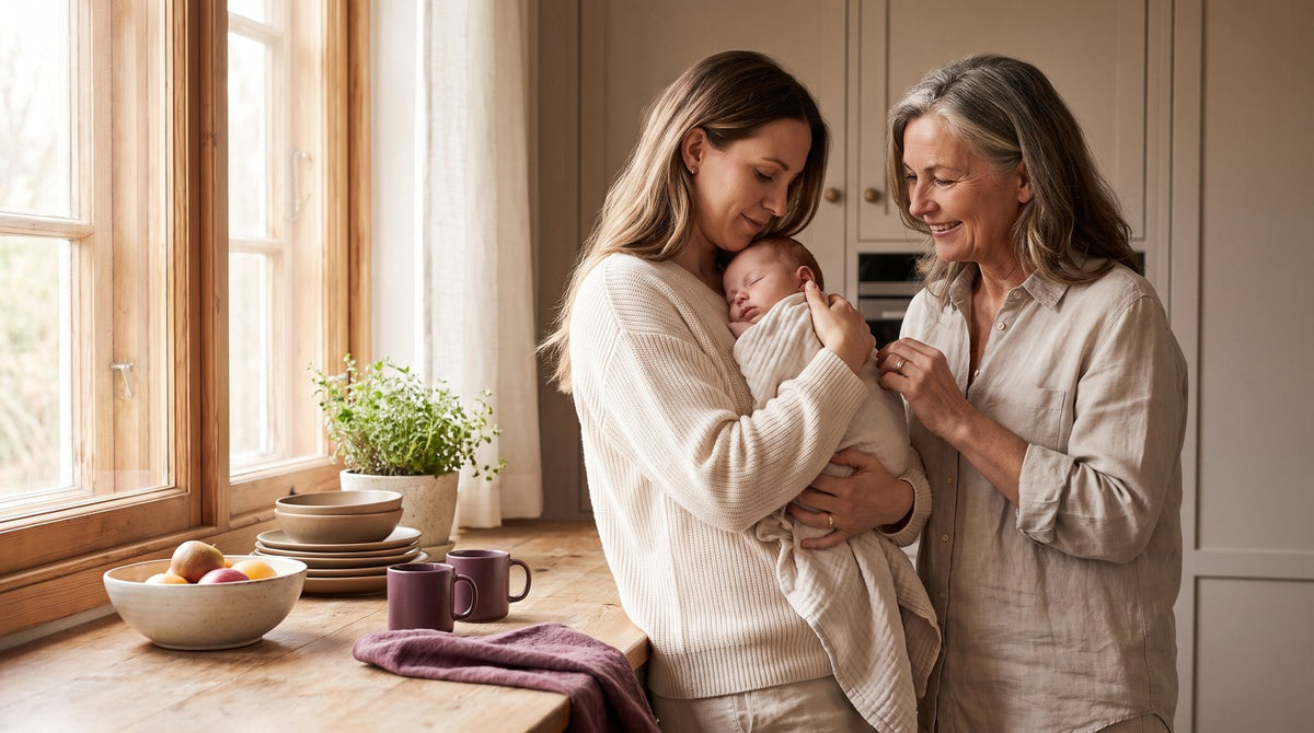 Young mother holding a sleeping baby beside an older woman in a warm kitchen