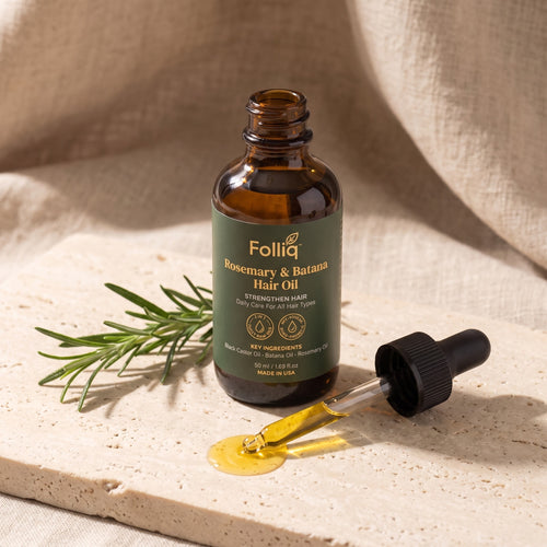 Folliq Rosemary & Batana Hair Oil bottle with its dropper, a drop of oil and a rosemary sprig on stone