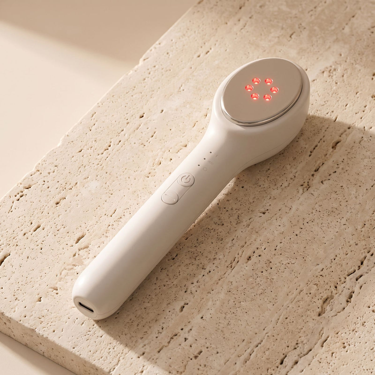 Folliq LED Face Wand lying on travertine stone, red 633 nm LEDs on, USB-C port at the base