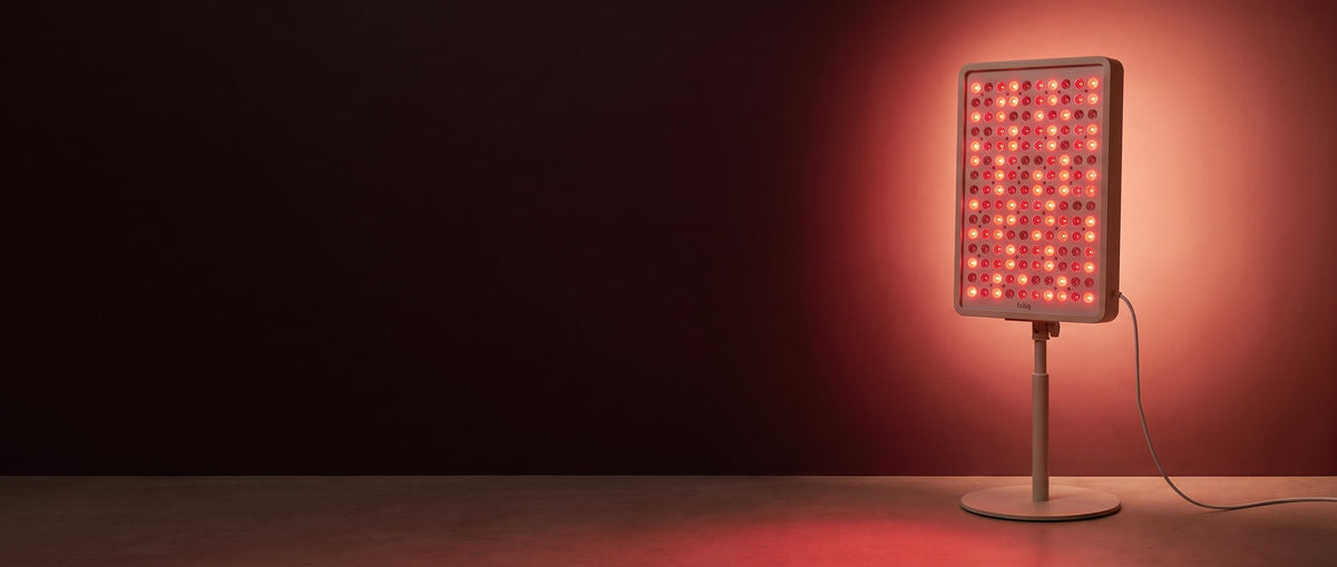 Folliq Red Light Therapy Panel in oat white on its floor stand, switched on and glowing red in a dark room
